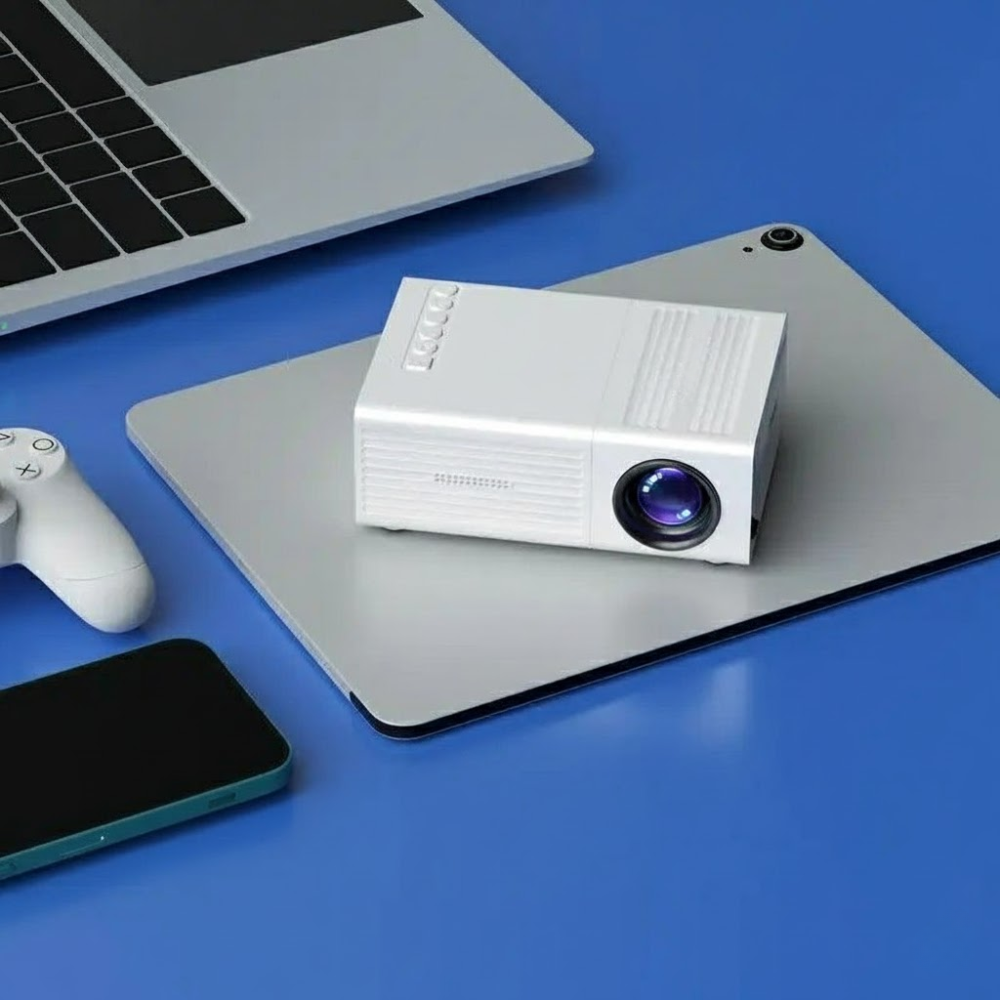 White projector on a blue surface with a laptop, smartphone, and gaming controller.