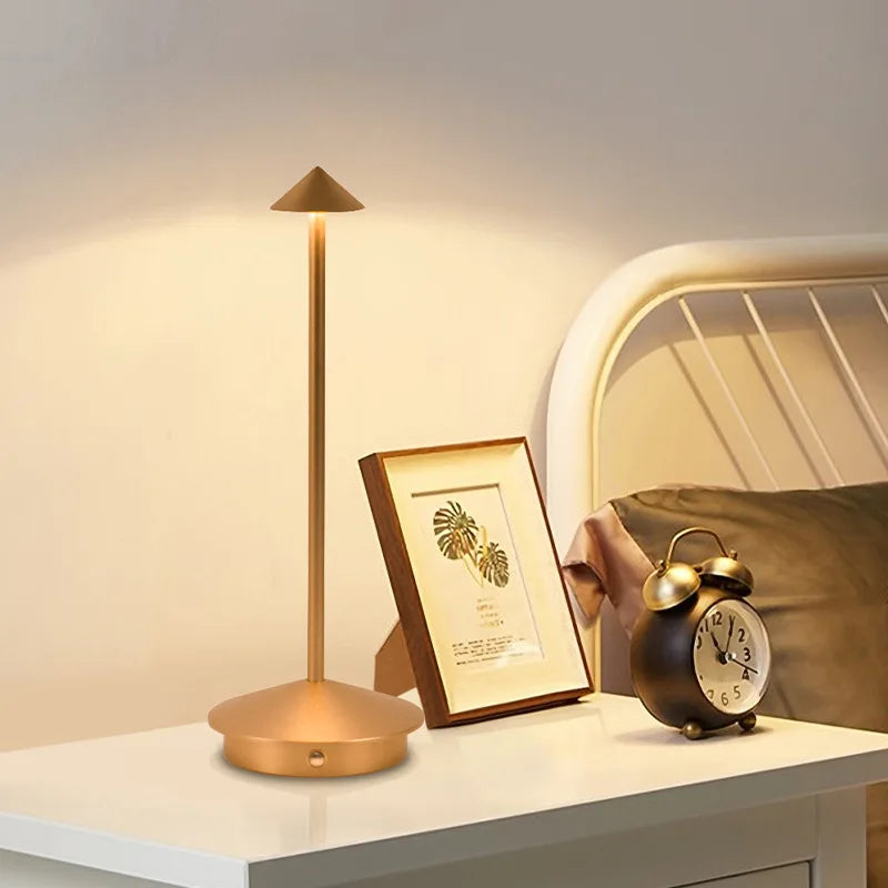 Gold lamp, photo frame, and alarm clock on a nightstand with a bed in the background