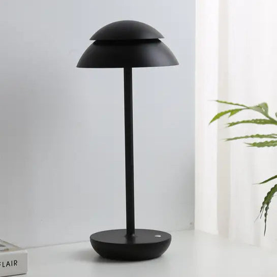 Black table lamp on a white surface with a light gray wall and plant in the background