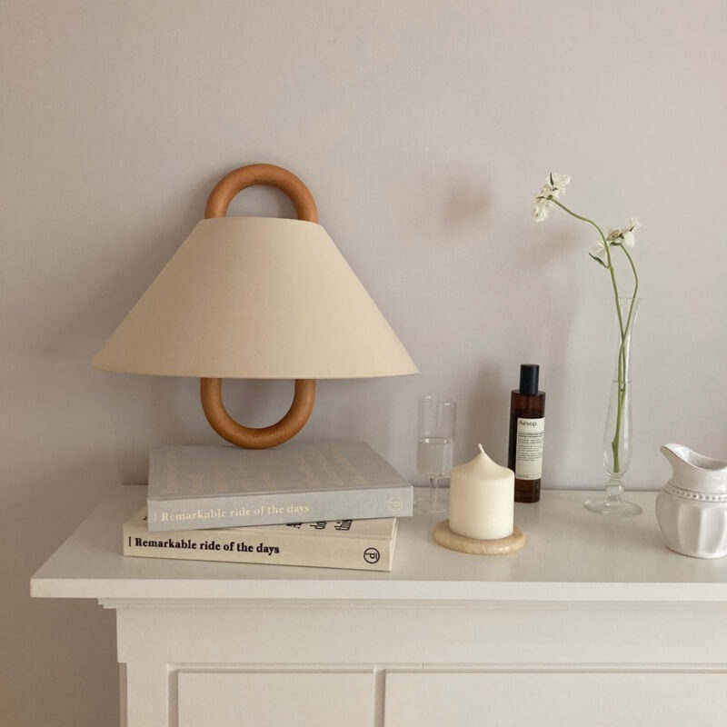 Table lamp with wooden base and beige shade on a white surface with books and decorative items.