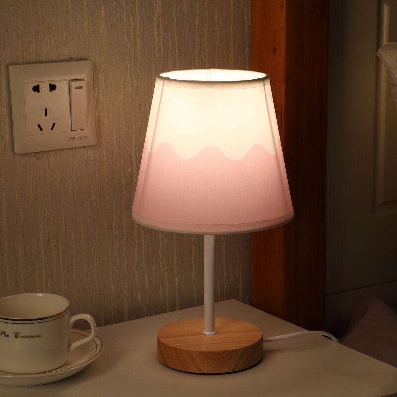Table lamp with a wooden base and pink gradient shade on a surface next to a cup and saucer.