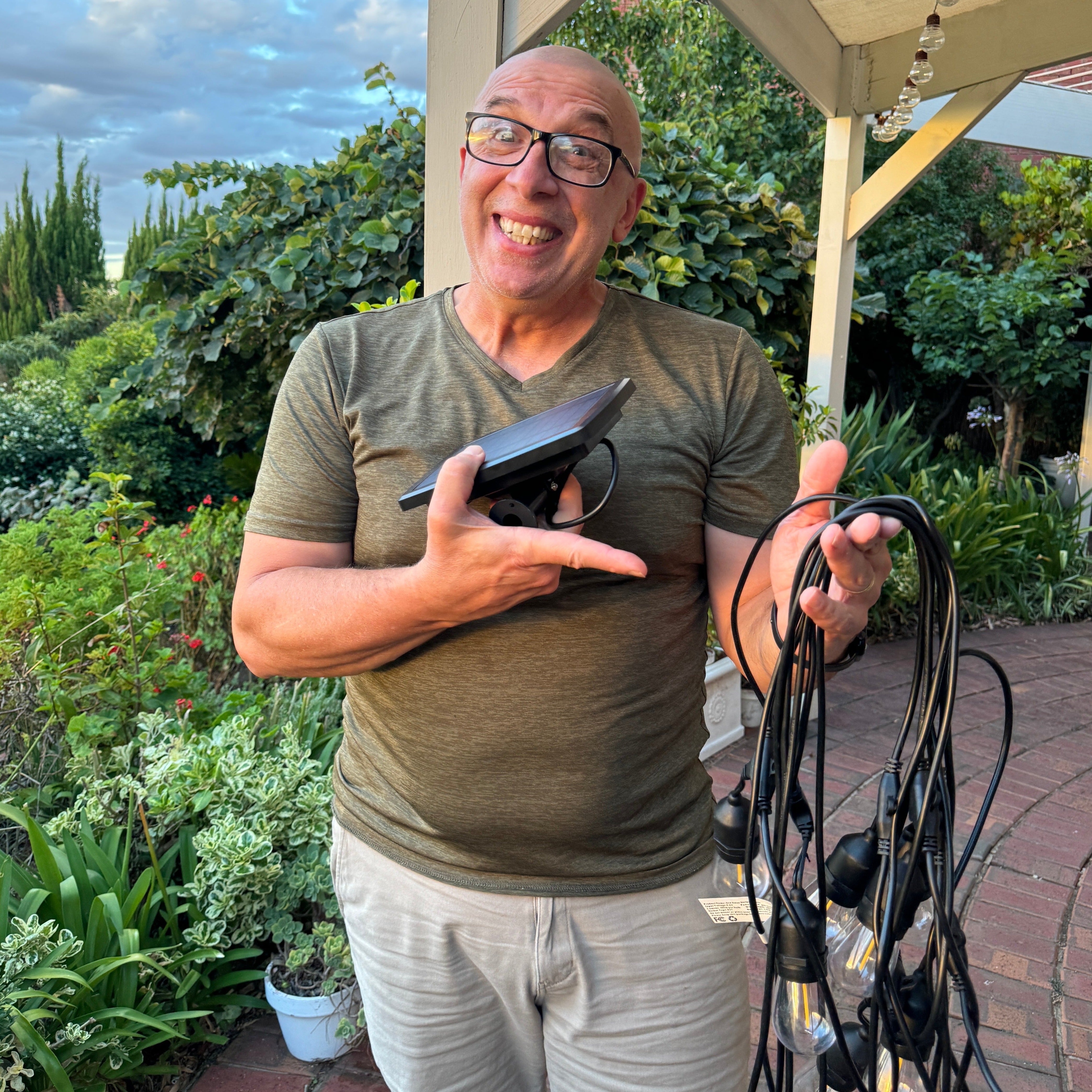 Man holding a string of lights outdoors with greenery in the background