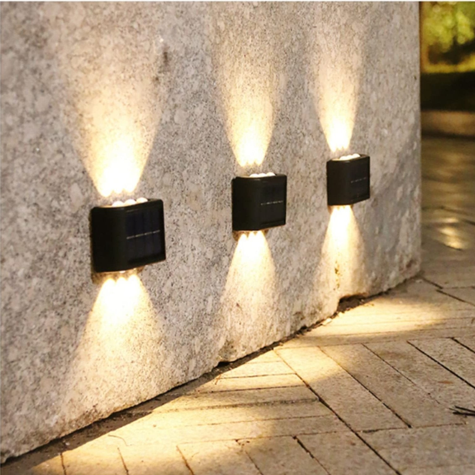 Three solar wall lights on a stone wall casting light on a pavement.