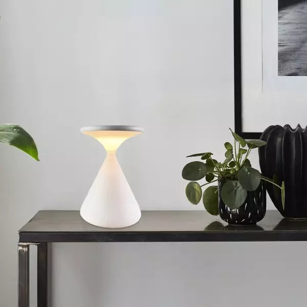 White table lamp on a dark surface with a plant and framed picture in the background