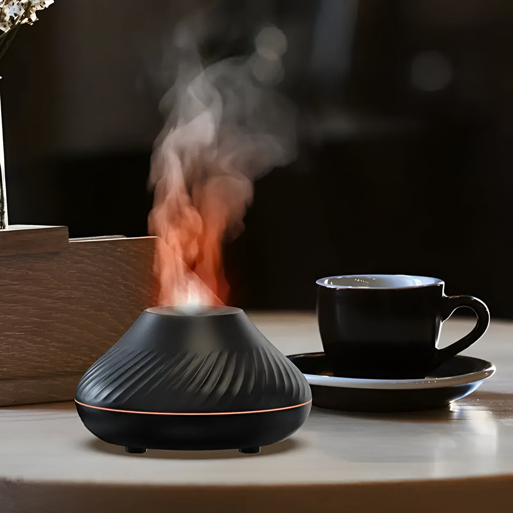 Black essential oil diffuser with steam emitting next to a black cup on a wooden surface.