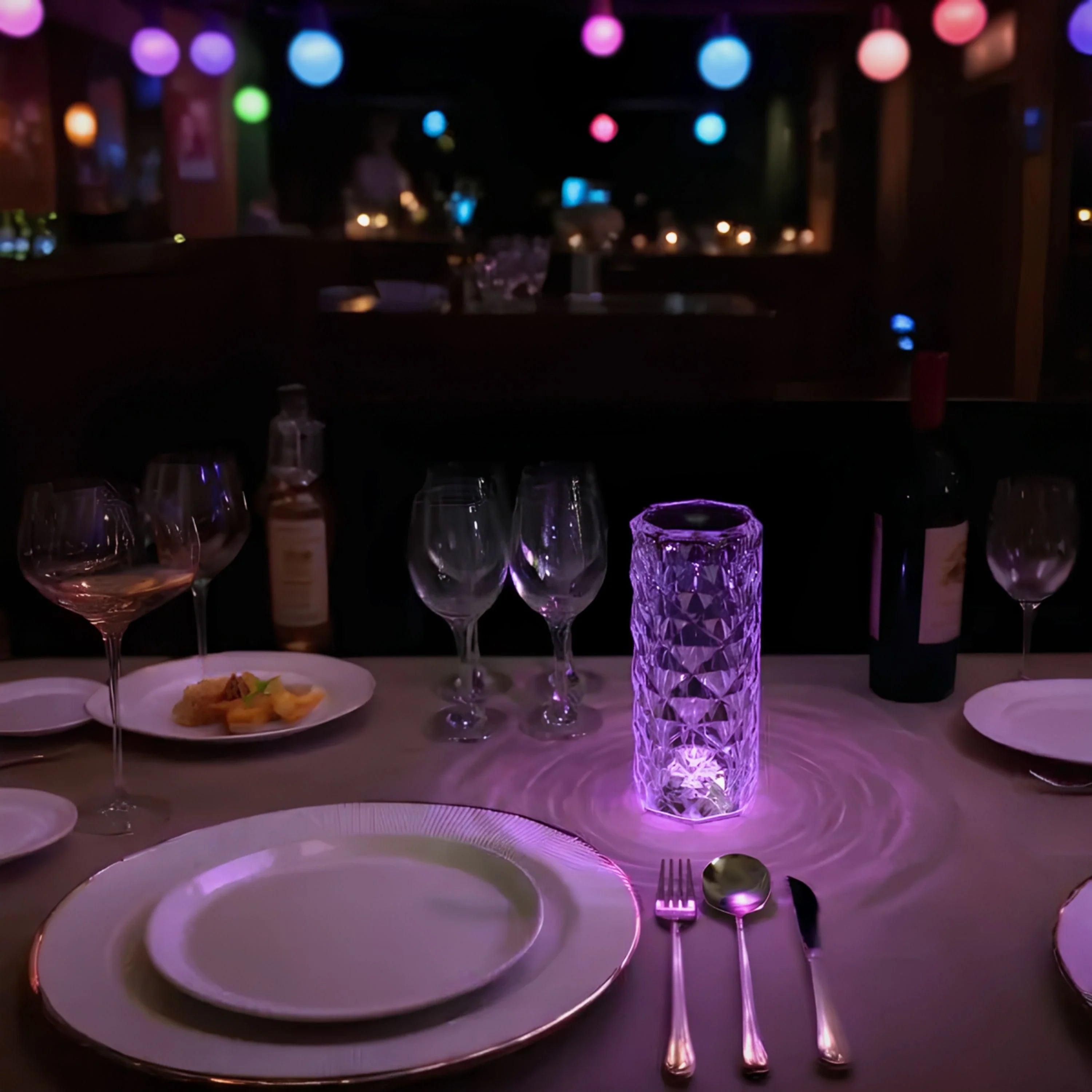 Dining table setup with plates, glasses, and cutlery in a dimly lit setting with colorful lights in the background.