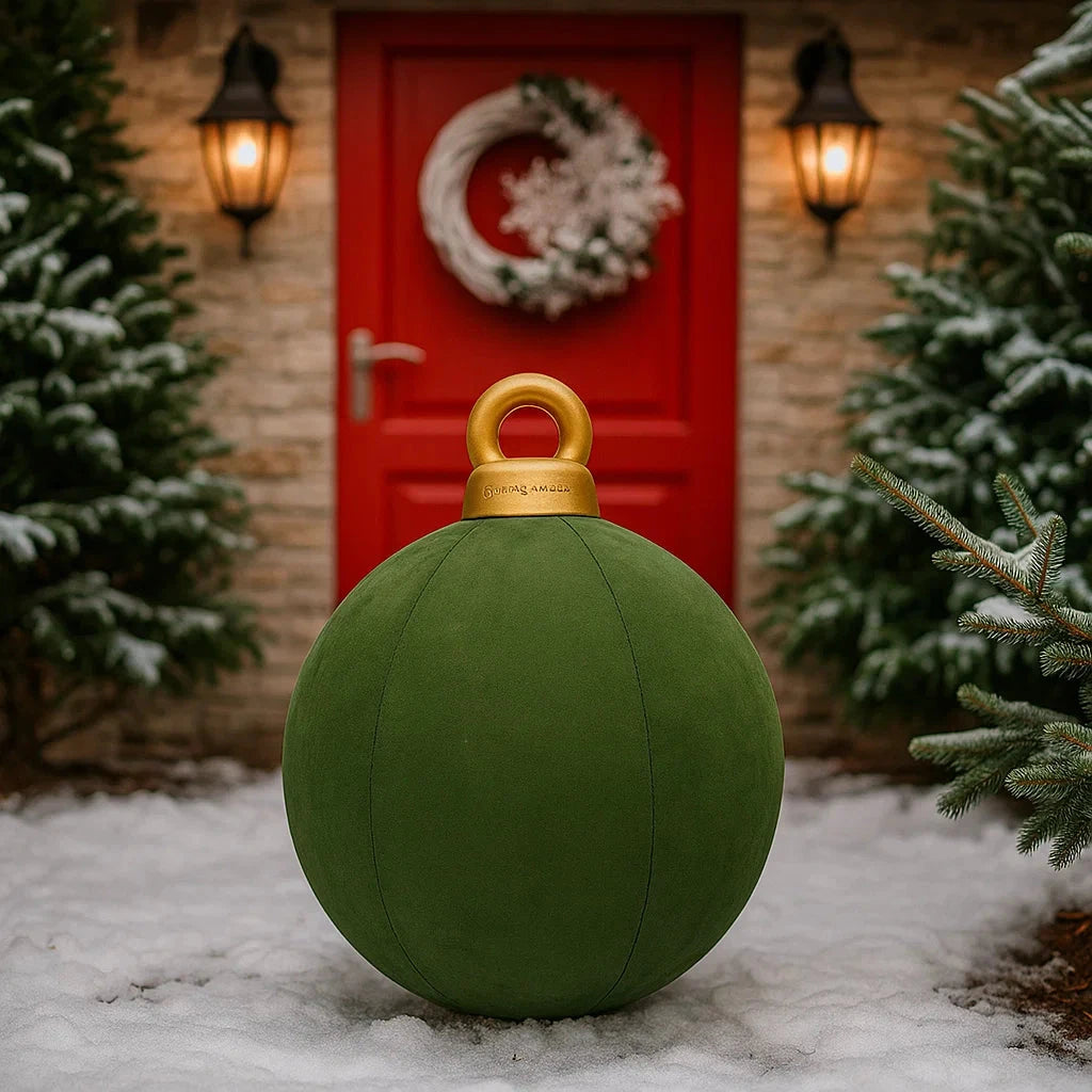 Large green Christmas ornament with gold top in front of a red door with a wreath, surrounded by snow and trees.