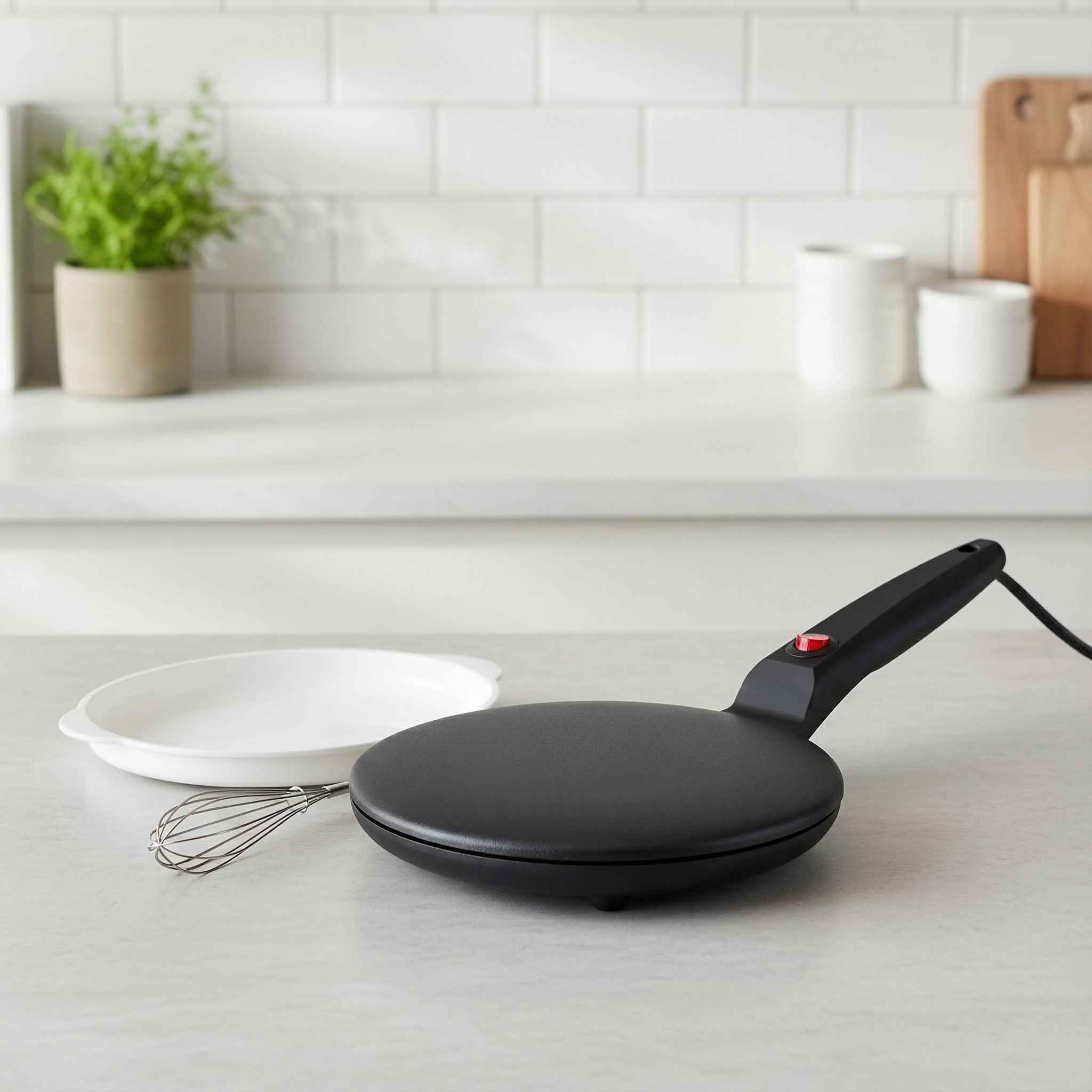 Black electric crepe maker on a kitchen counter with a whisk and plates in the background.
