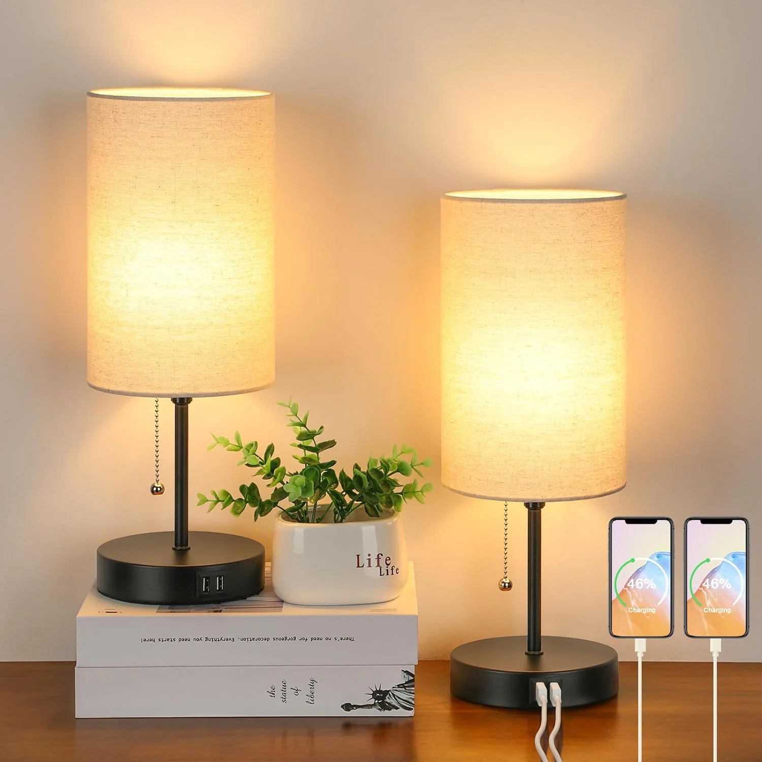 Two table lamps with fabric shades on a wooden surface, one with a plant and books, the other with a smartphone.