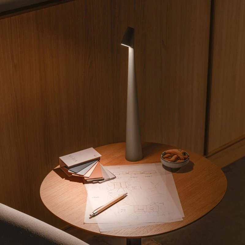 Modern table lamp on a wooden table with papers and a pen in a dimly lit room.