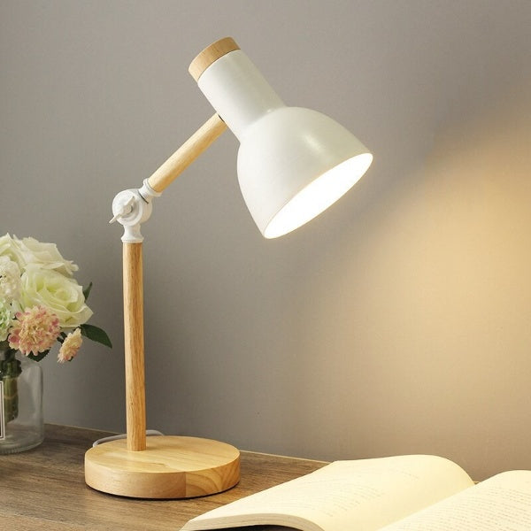 Table lamp with wooden base and white shade on a desk with flowers and book.