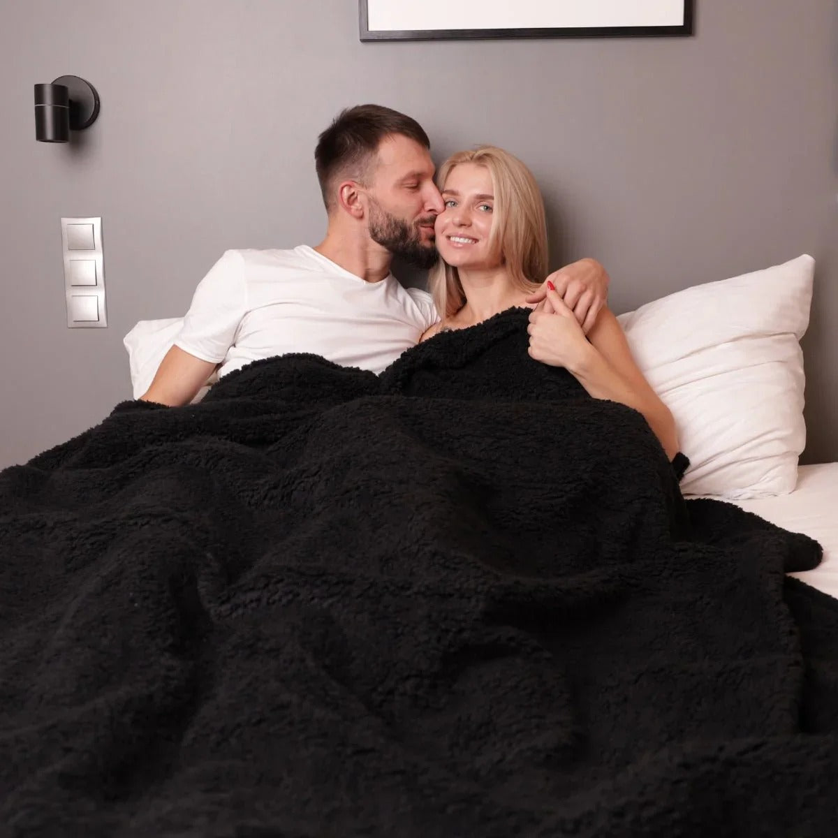 Couple under a black blanket on a bed in a bedroom setting.