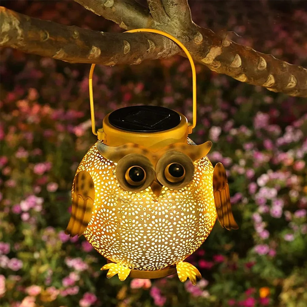 Owl-shaped solar lantern hanging on a branch with a blurred floral background