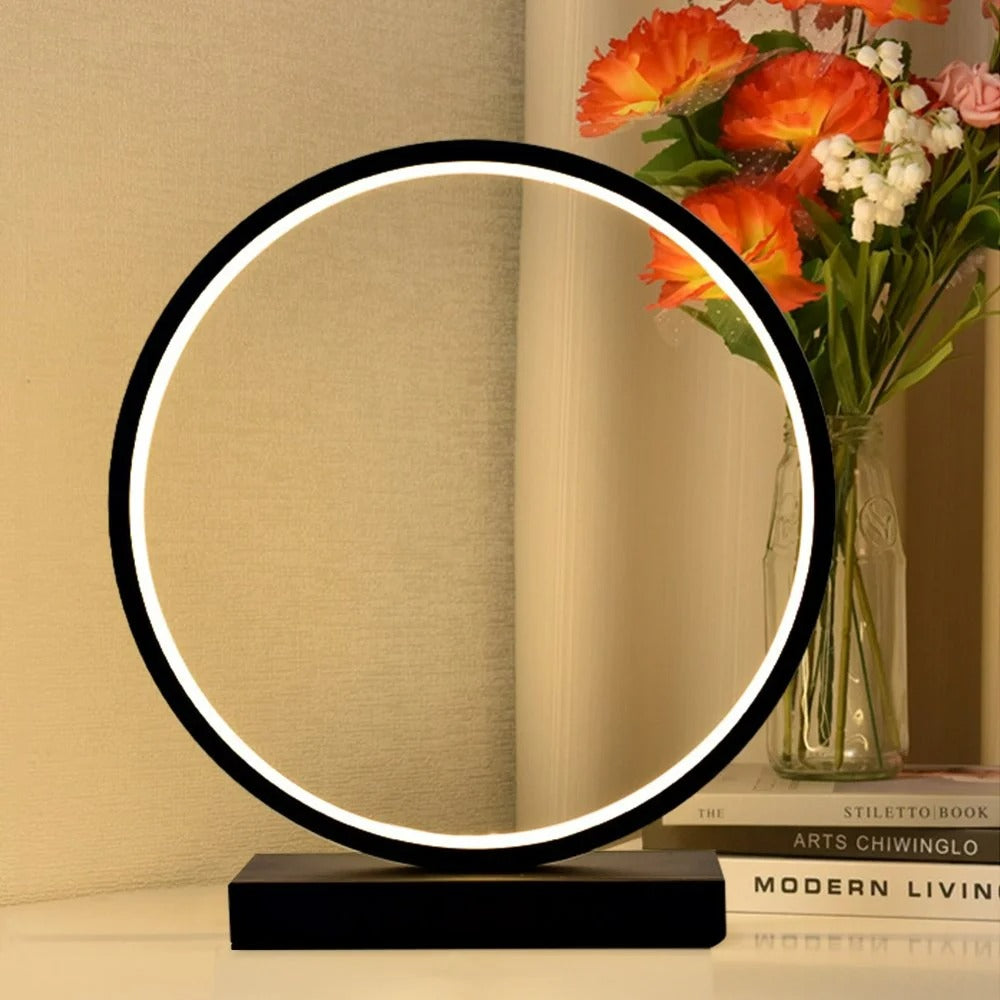 Modern table lamp with circular design on a wooden surface, next to a vase of flowers.