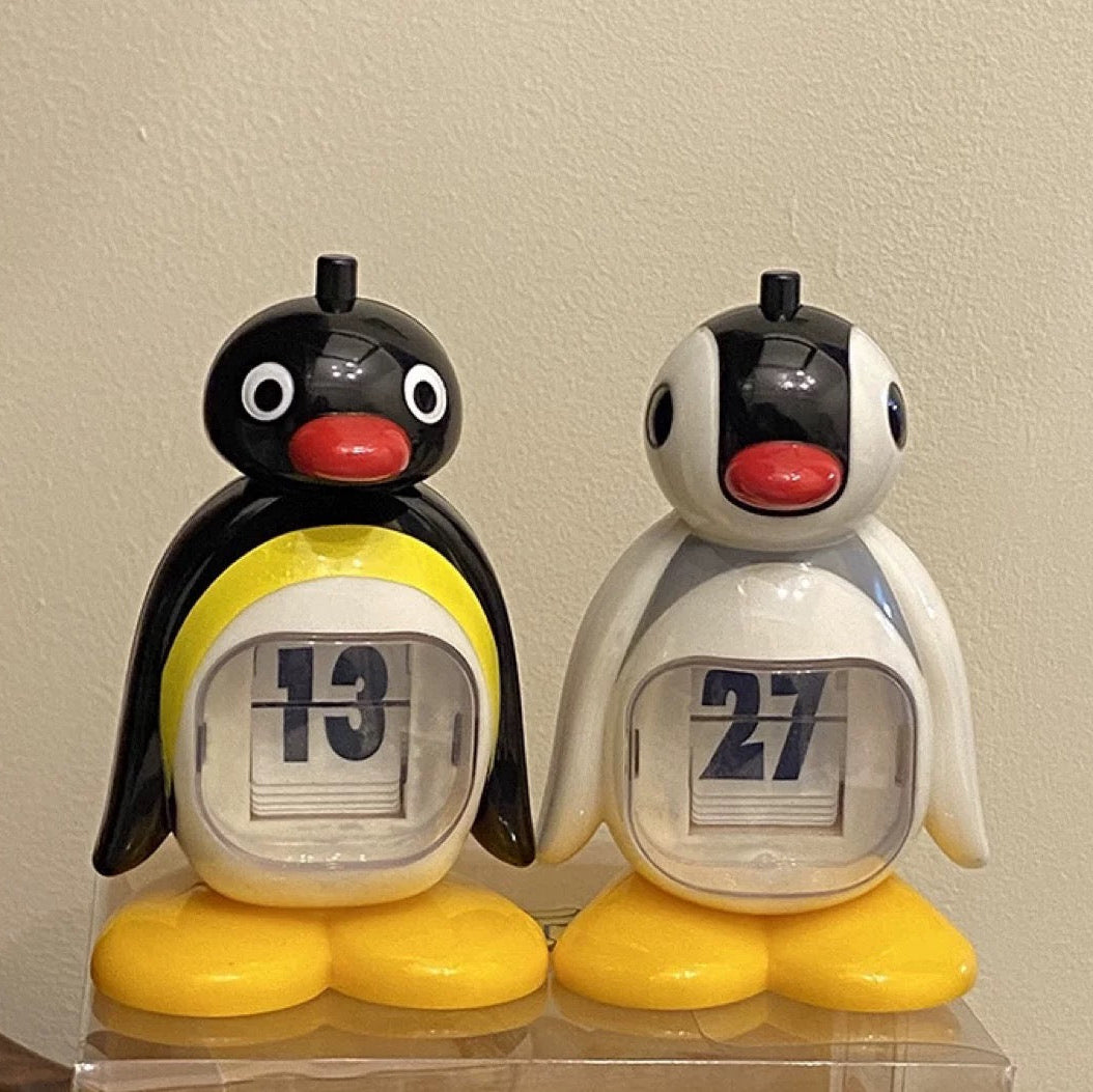 Two penguin-shaped calendars on a clear stand against a beige wall.