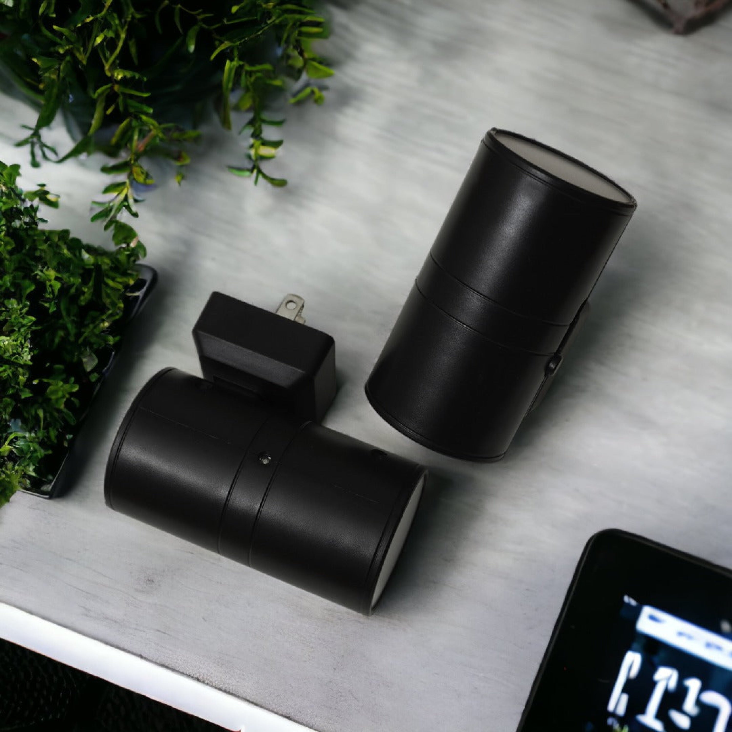 Three black cylindrical devices on a gray surface with a smartphone displaying time.