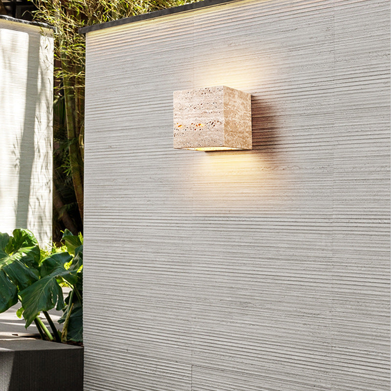 Square outdoor wall light fixture on a textured wall with plants in the background