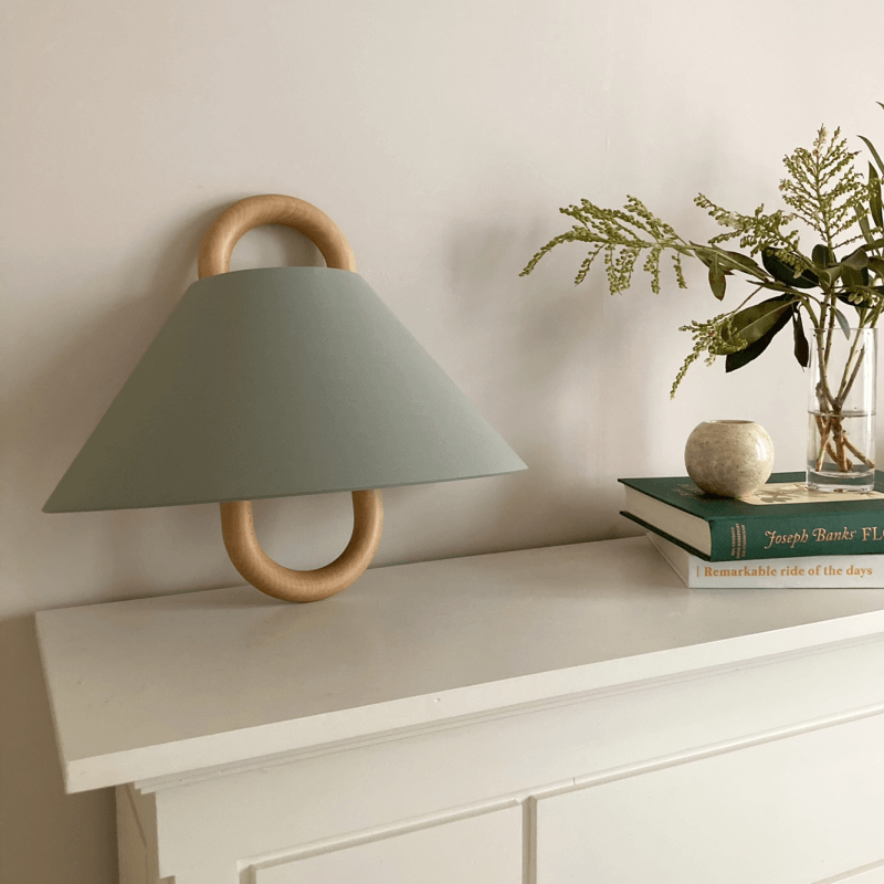 Modern wall lamp with wooden accents on a white surface next to books and a plant.