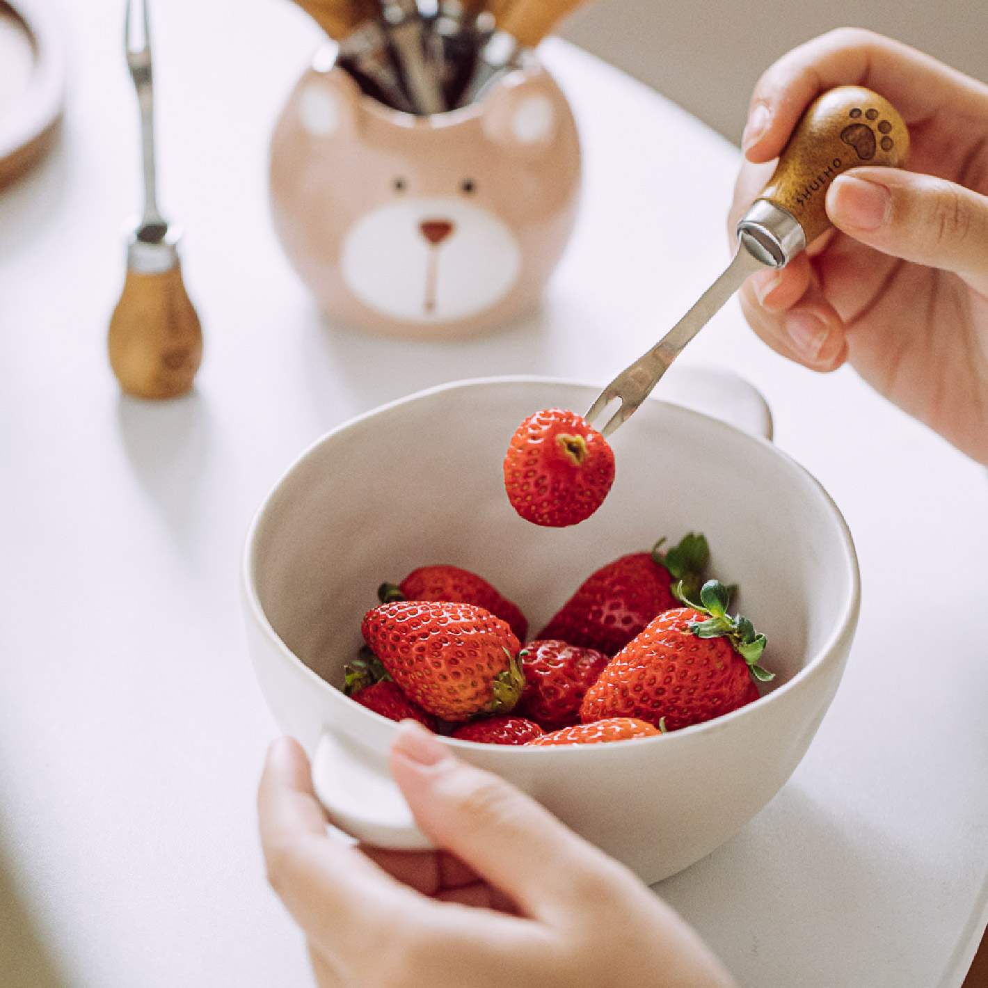 Person picking a strawberry from a bowl with a bear-shaped container in the background