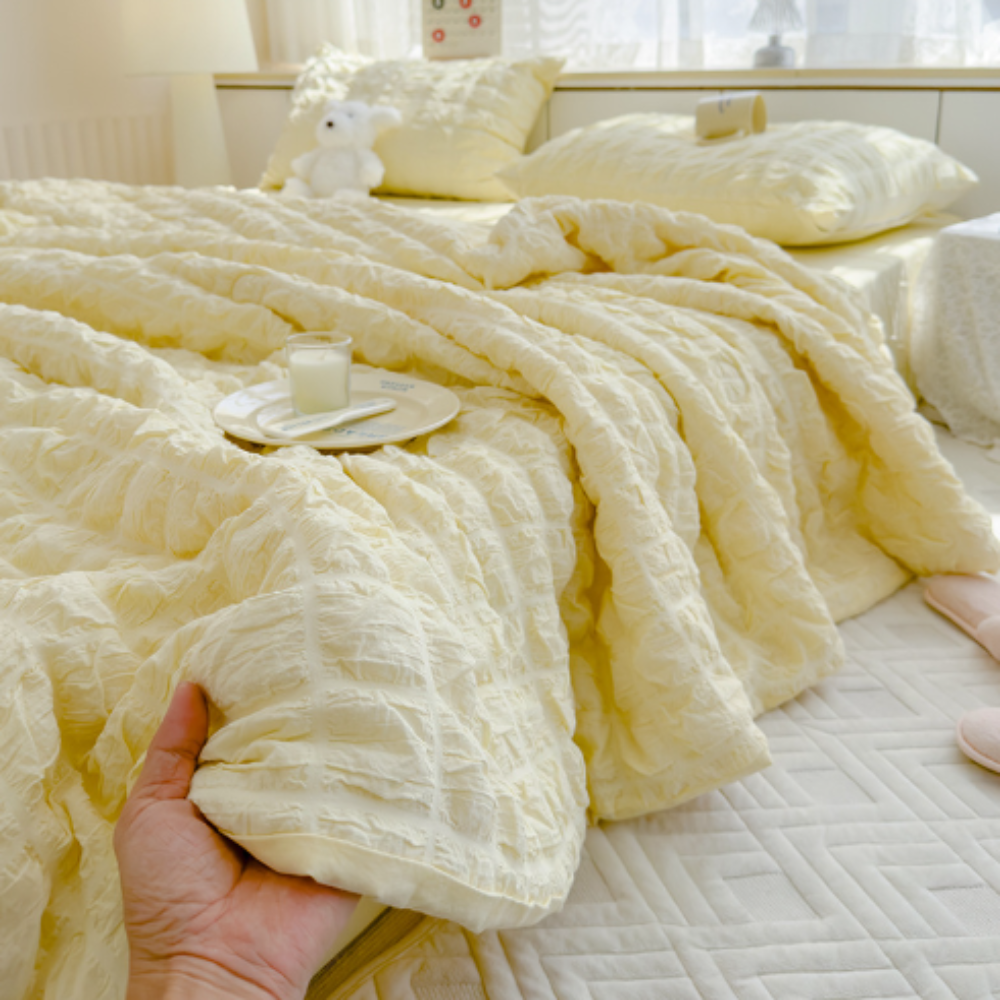 Hand holding a textured yellow blanket over a bed with pillows and a small table.