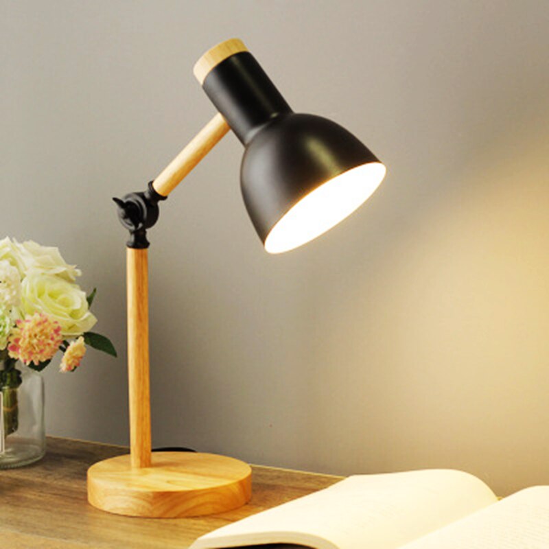 Table lamp with black shade and wooden base on a desk with flowers and book.