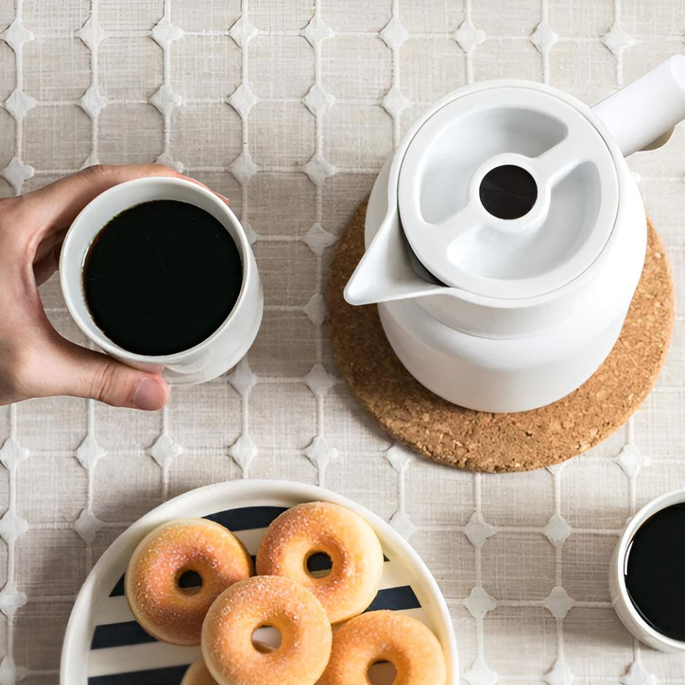 Person holding a cup of coffee next to a white teapot and plate of donuts on a patterned surface.