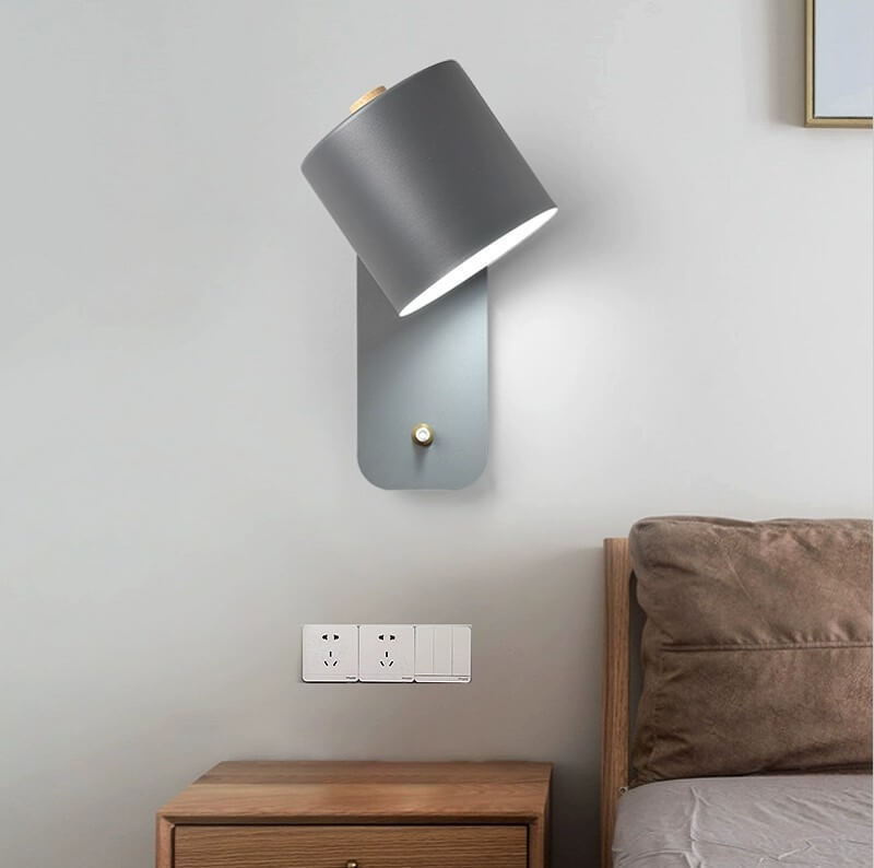 Gray wall lamp on a light gray wall above a wooden nightstand with a bed in the background.