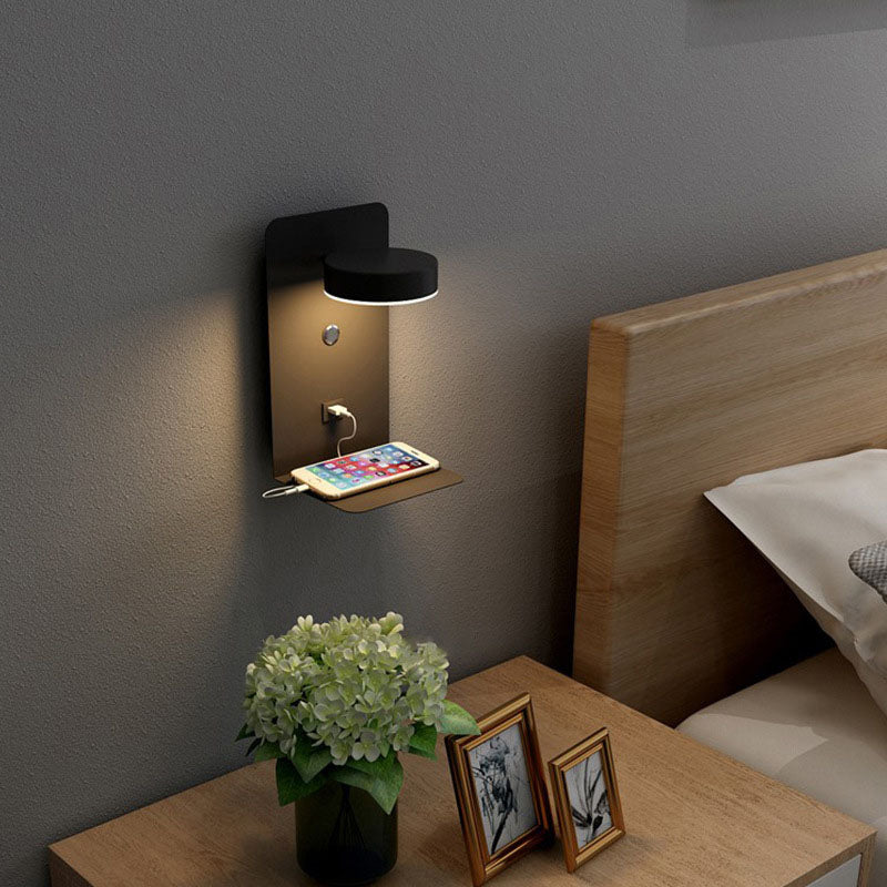 Wall-mounted night light with phone charging station on a bedside table next to a bed.