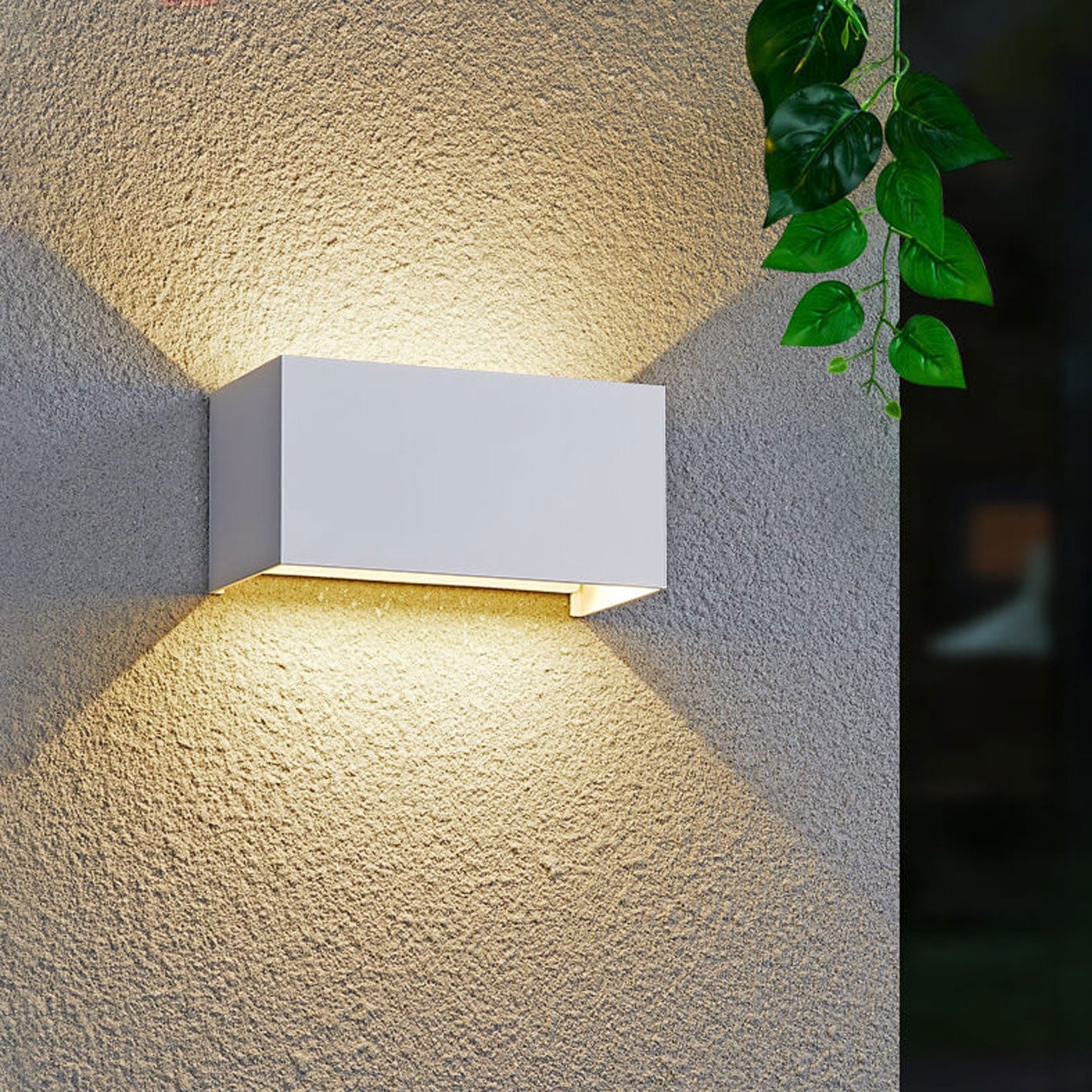 White outdoor wall light fixture on a textured wall with a plant in the background