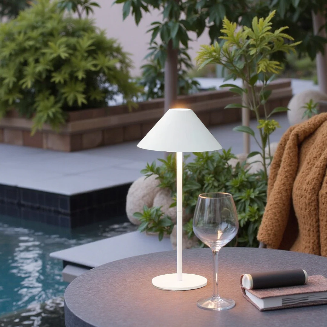 White table lamp on a table by a pool with plants and a glass in the background