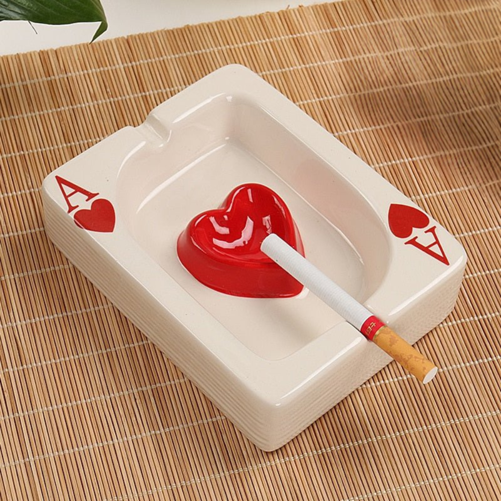 White ashtray with red heart design and 'Ace of Hearts' smoking accessory on a woven mat.