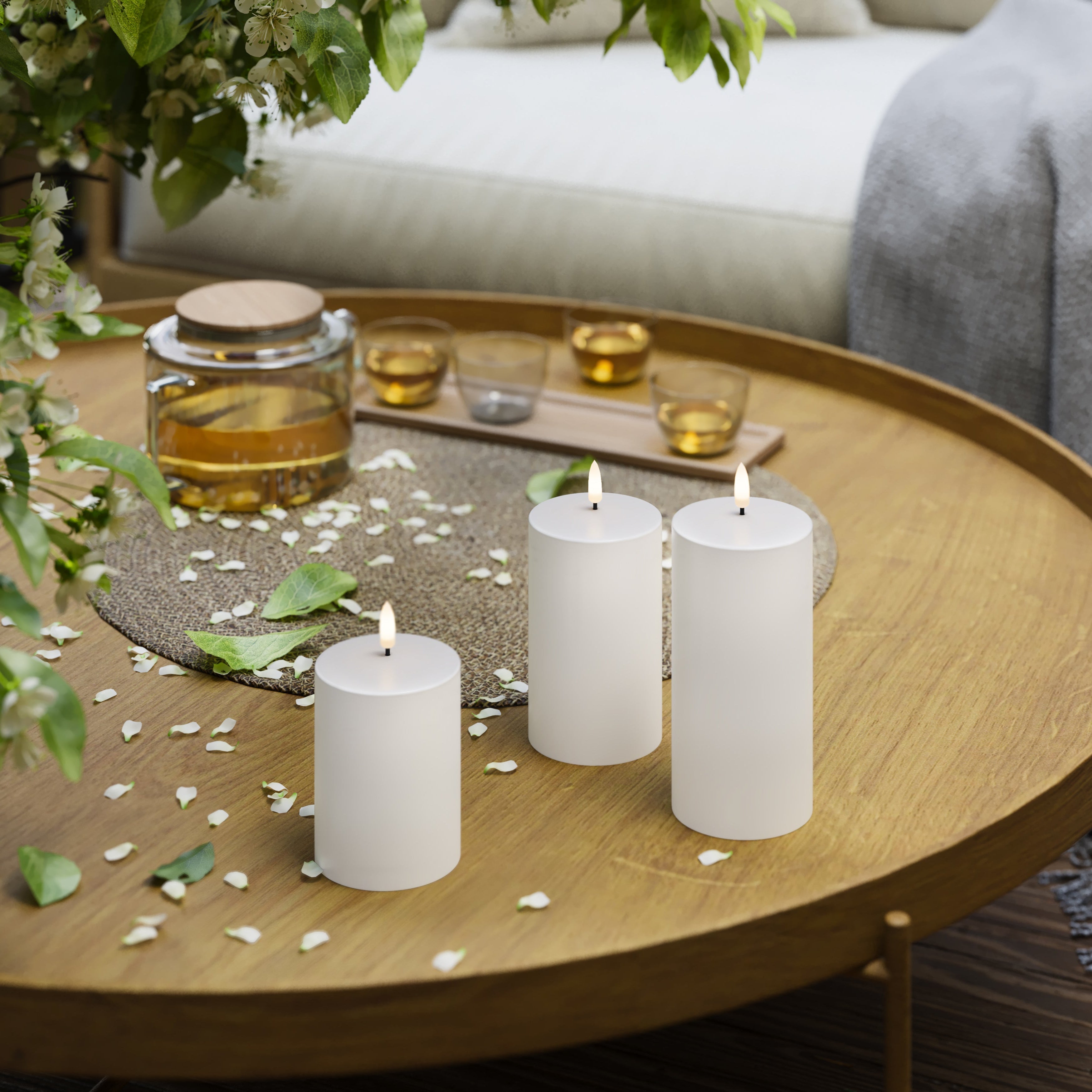Three lit candles on a wooden table with a teapot and cups in the background.