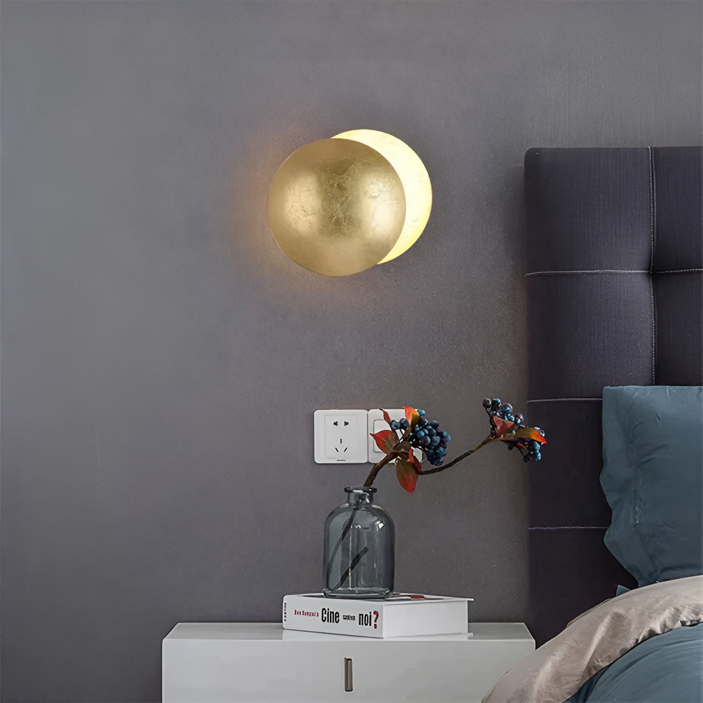 Gold spherical wall light fixture on a gray wall with a bed and nightstand in the foreground.