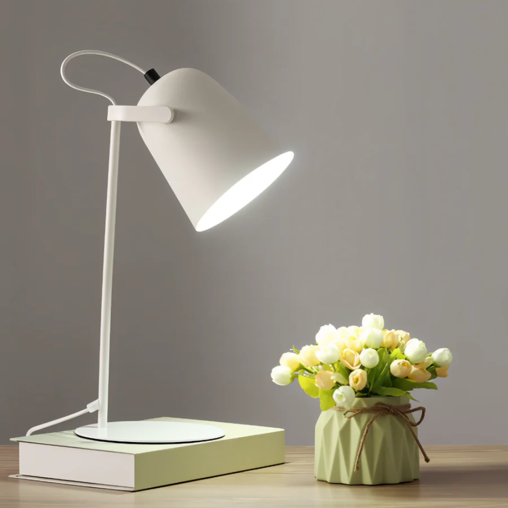 White desk lamp on a wooden surface with a small vase of flowers next to it.