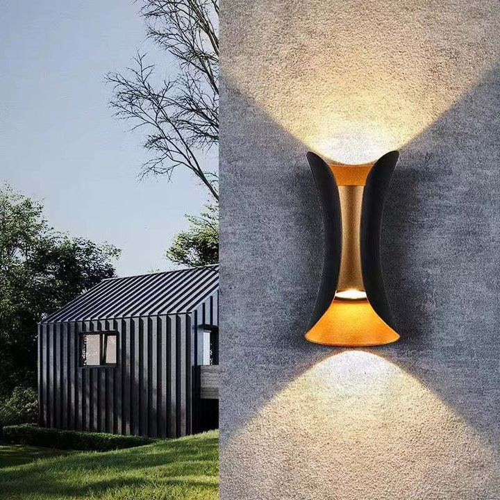Modern outdoor wall light fixture on a concrete wall with a house and trees in the background.