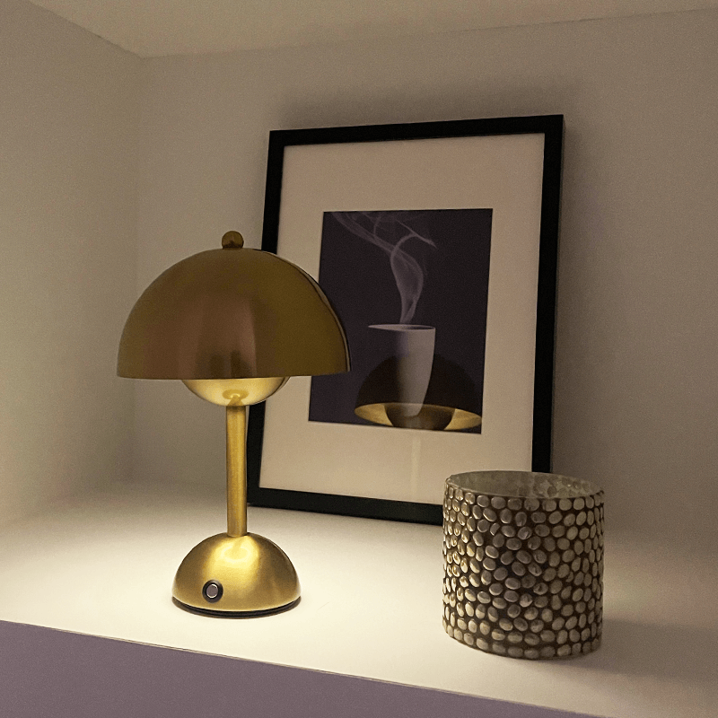 Gold table lamp on a surface with a framed picture and decorative pot in the background.
