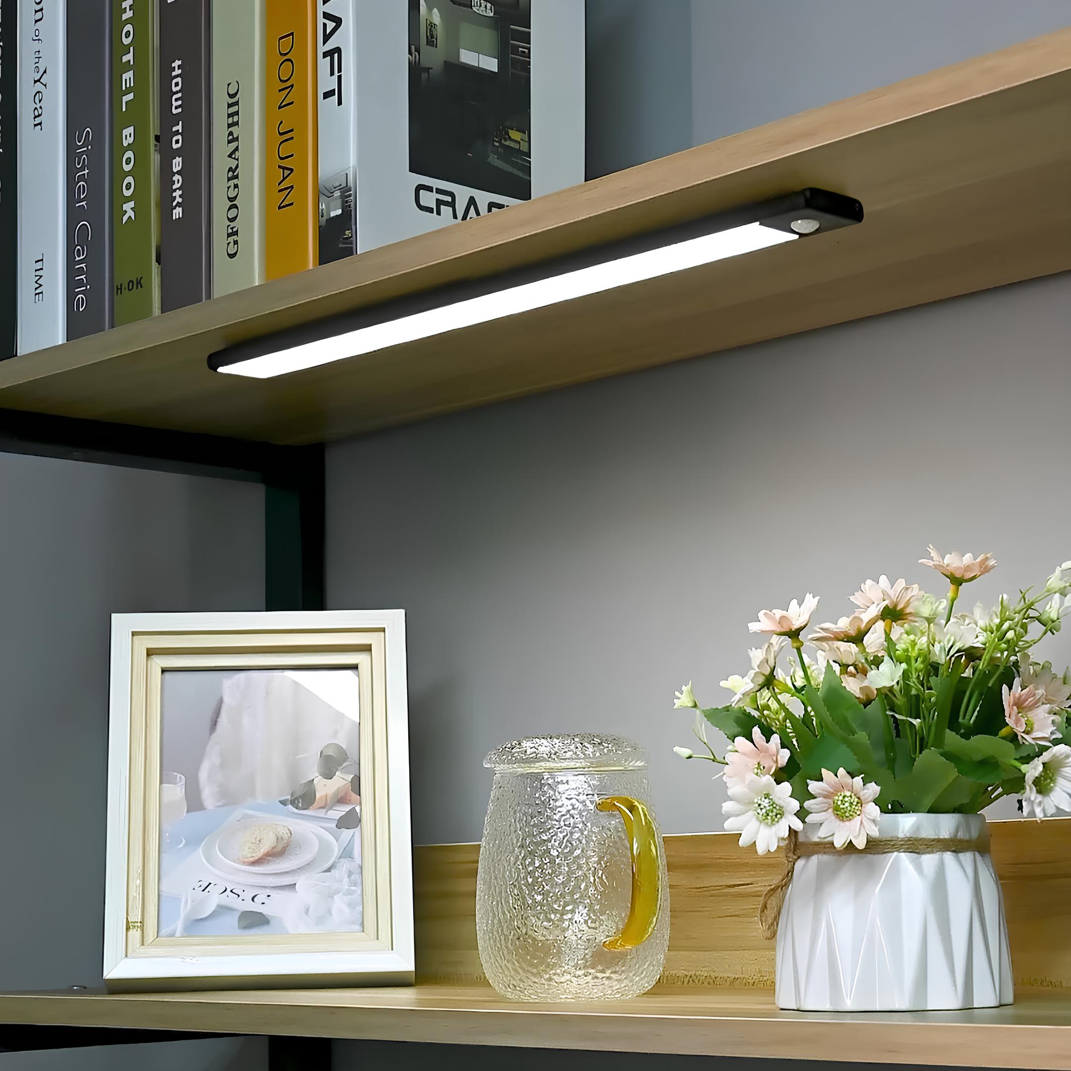 Wooden shelf with books, a picture frame, a glass pitcher, and a vase of flowers against a gray wall.