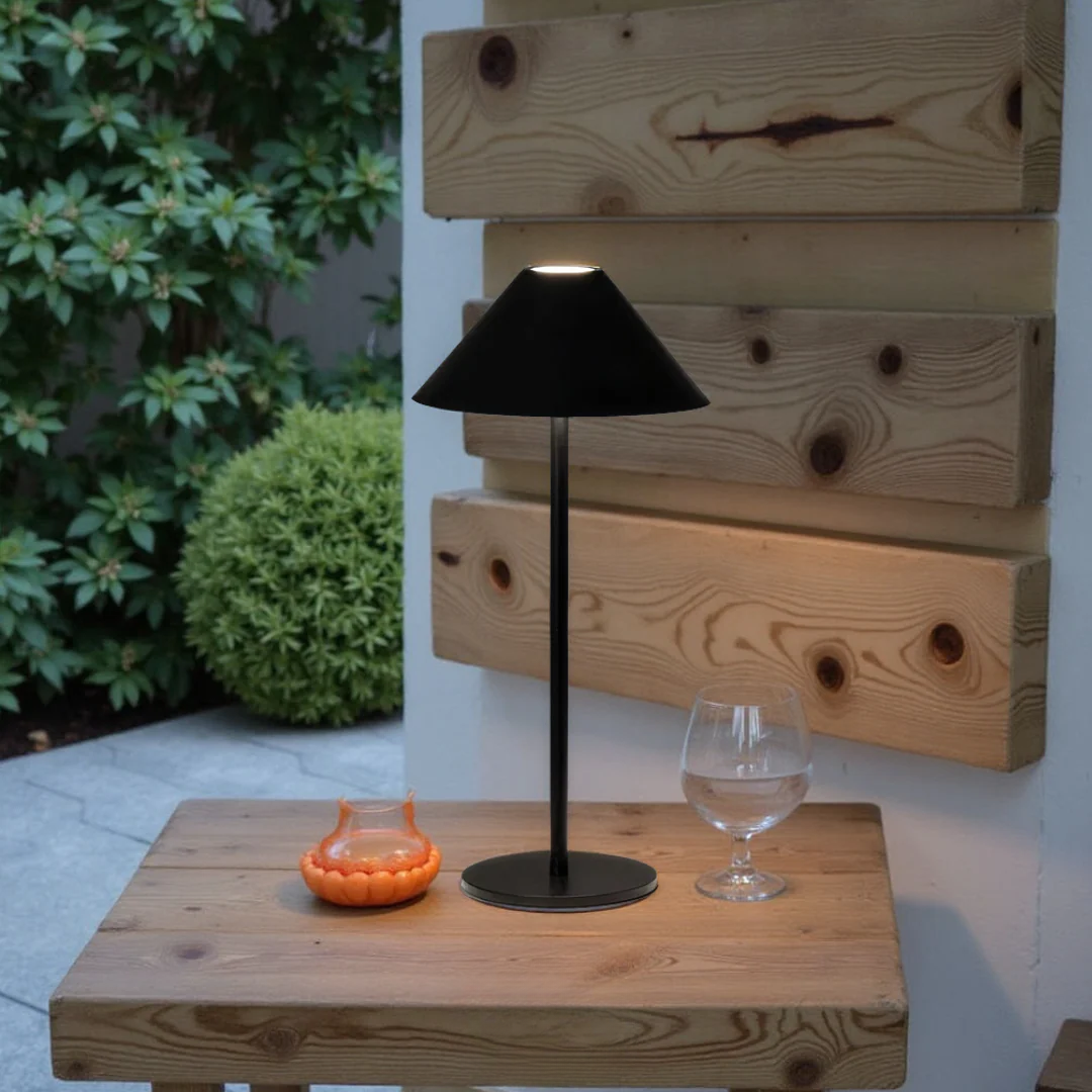 Black outdoor lamp on a wooden table with a glass and small orange object, against a wooden wall and greenery.