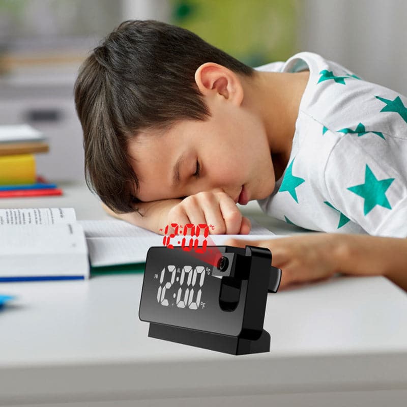 Child sleeping on a desk with an alarm clock showing 12:00.