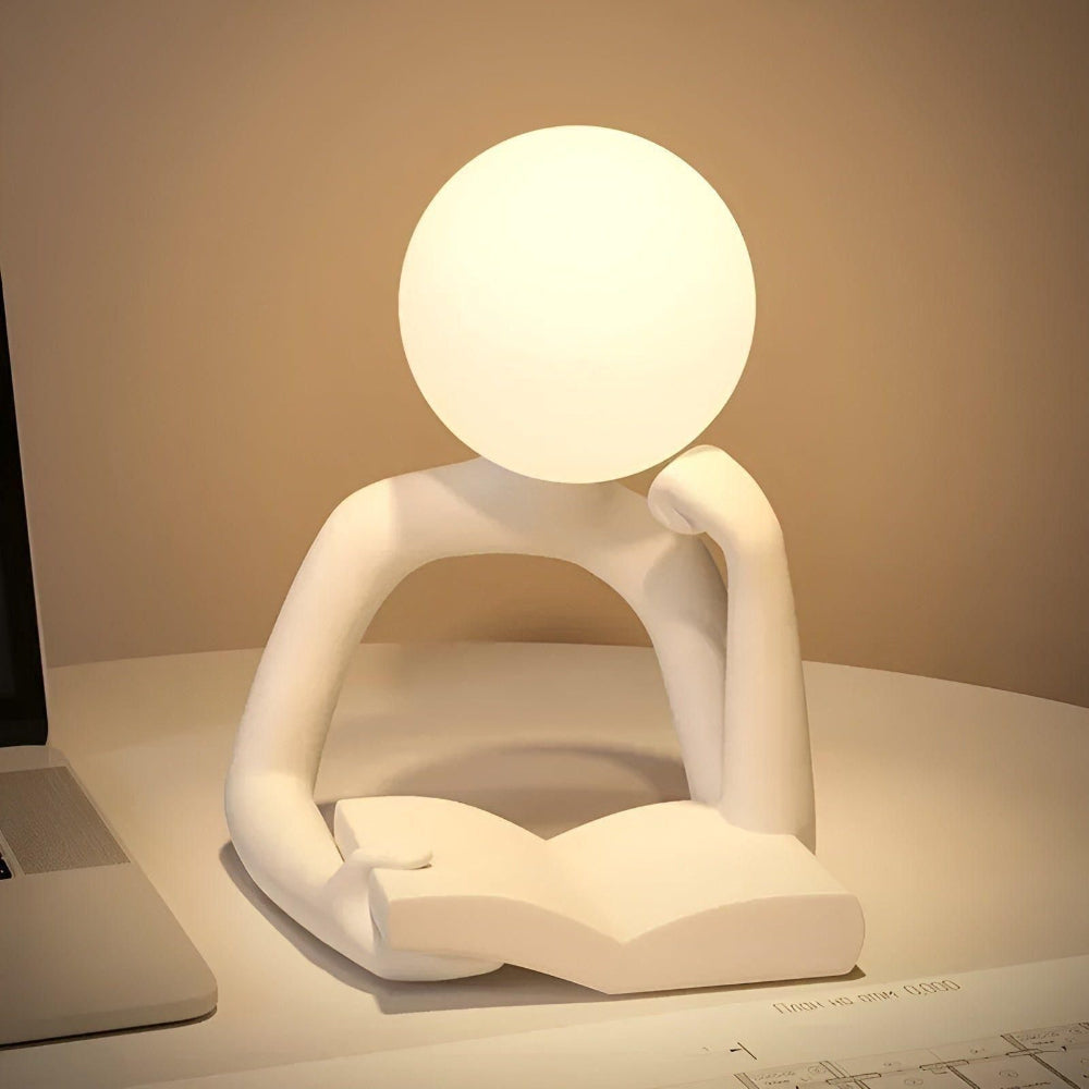 Sculptural lamp in the shape of a person reading a book on a desk.