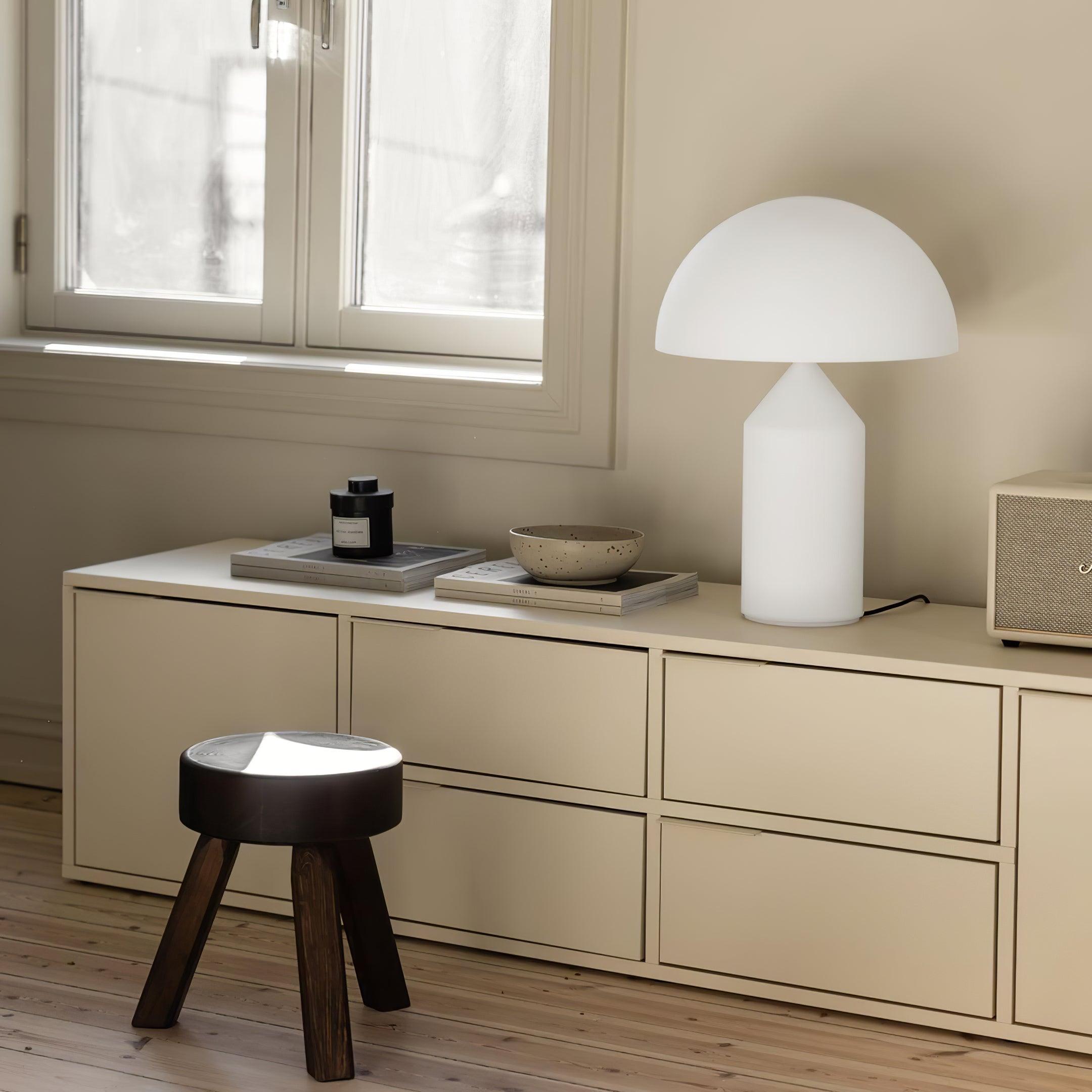 Modern interior with a white table lamp, wooden stool, and decorative items on a beige cabinet.
