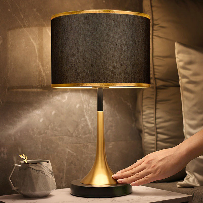 Table lamp with black shade and gold base on a marble surface, with a person's hand touching it.