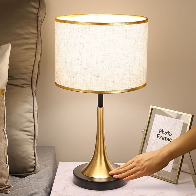 Table lamp with a gold base and white shade on a table next to a sofa and photo frame.