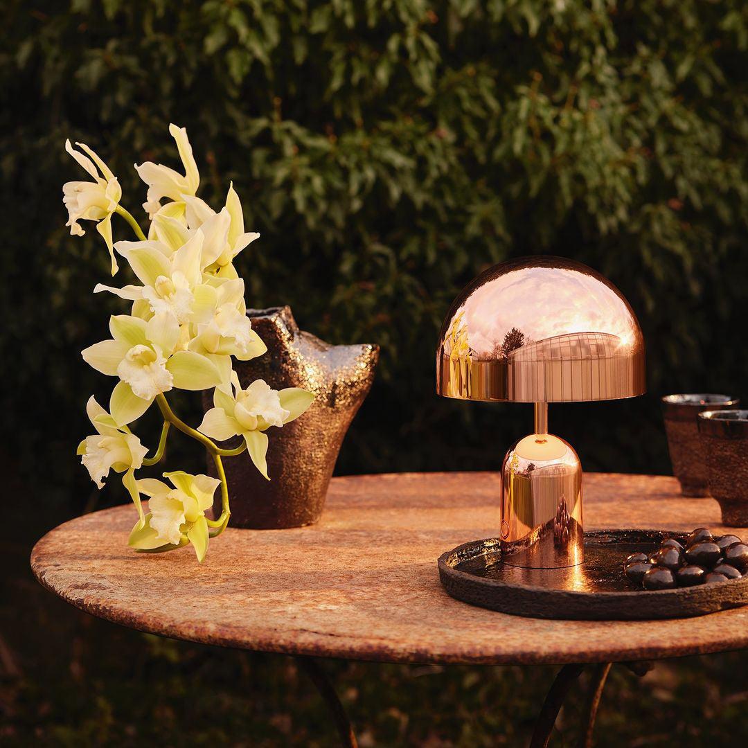 Gold table lamp on a wooden table with flowers and decorative items outdoors.