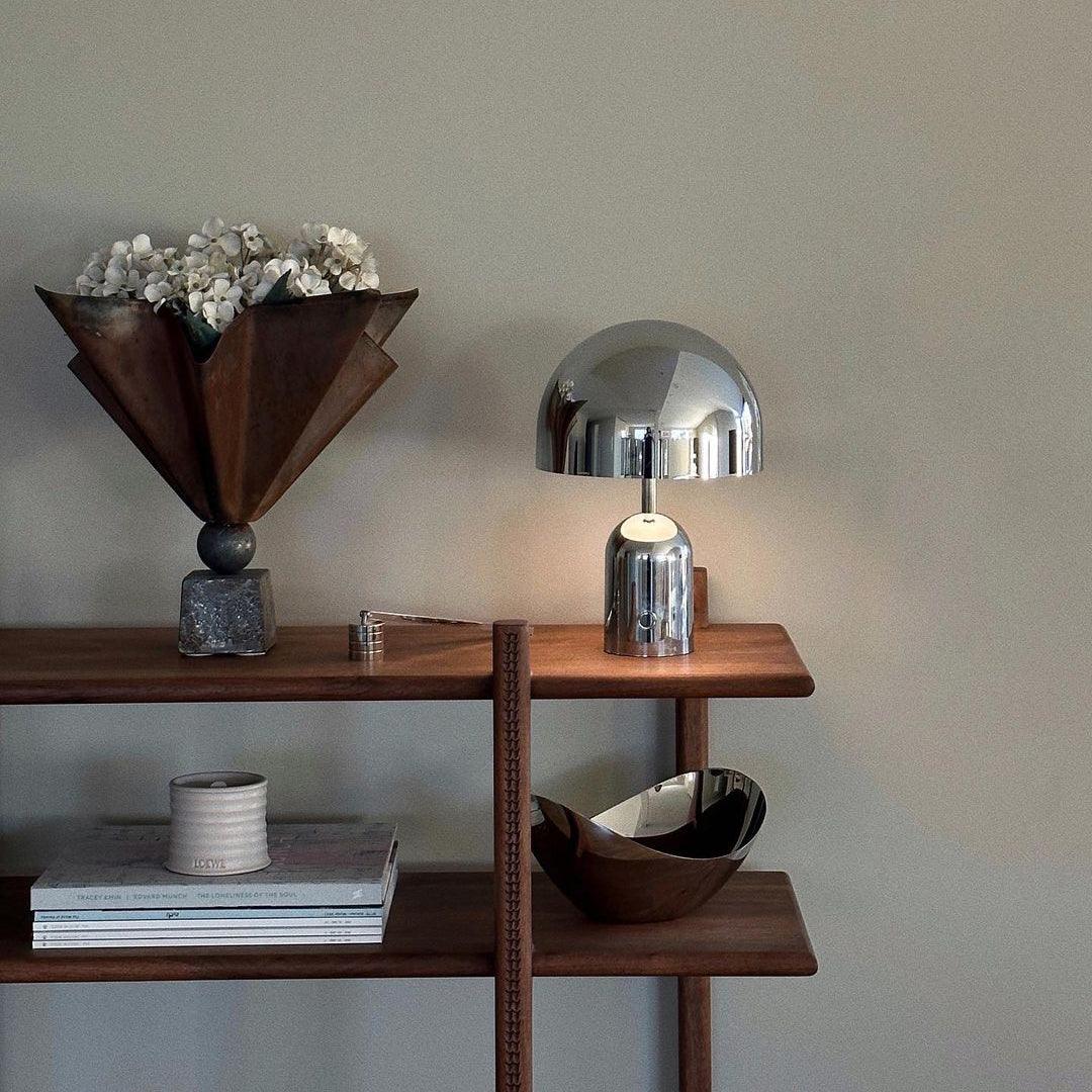 Wooden shelf with decorative items including a metallic lamp, vase, and books against a plain wall.
