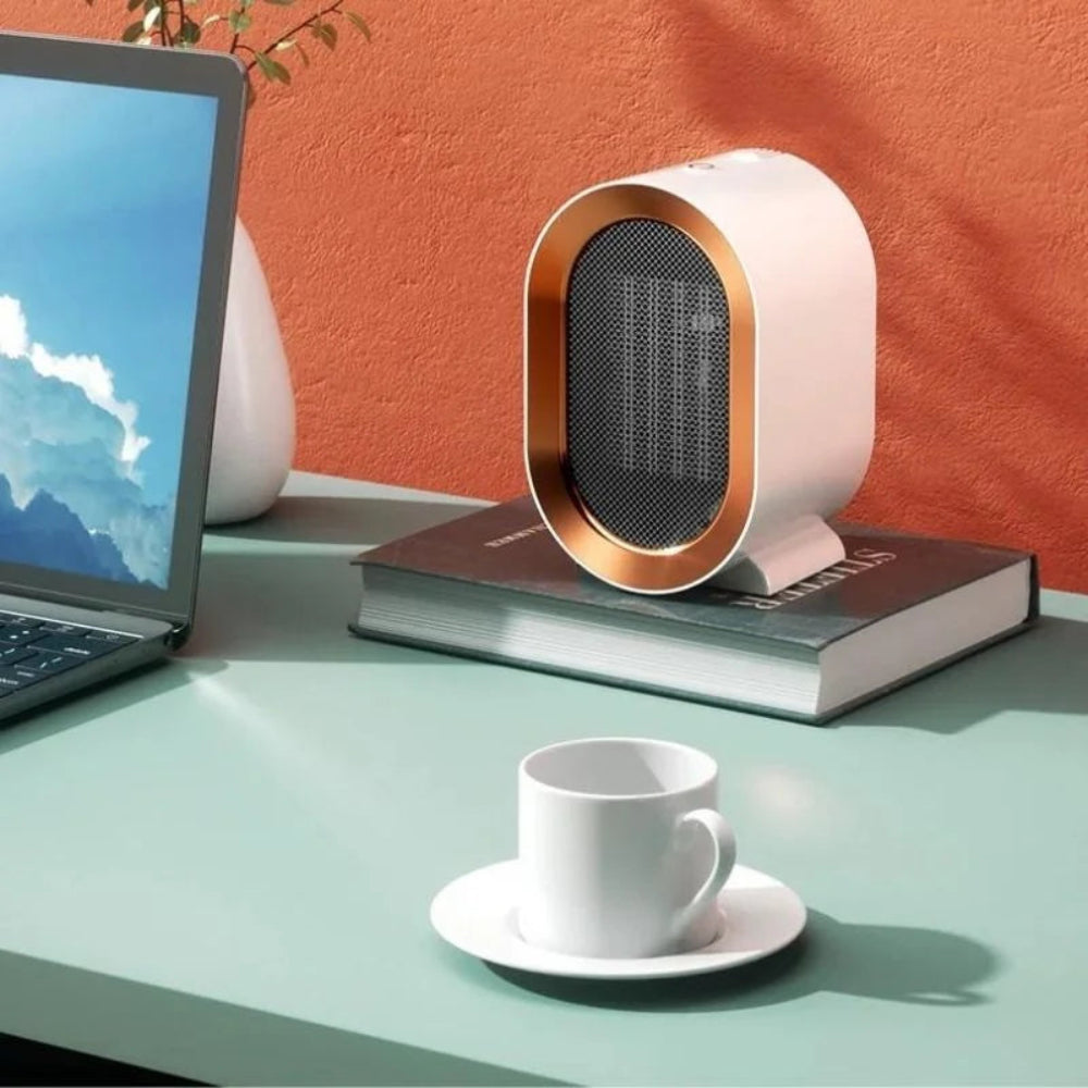 Small space heater on a book with a laptop and cup on a desk against an orange wall.