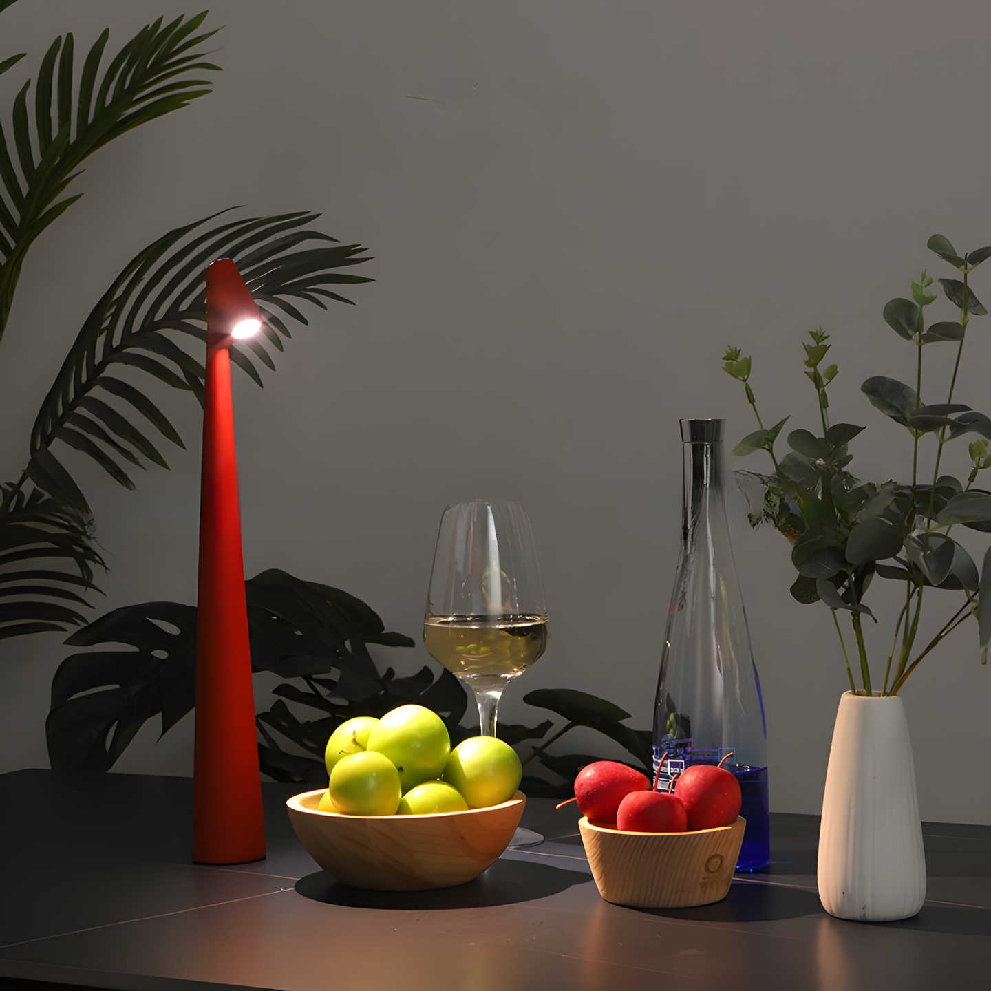Red candle-style lamp on a table with fruit, wine glass, and plant
