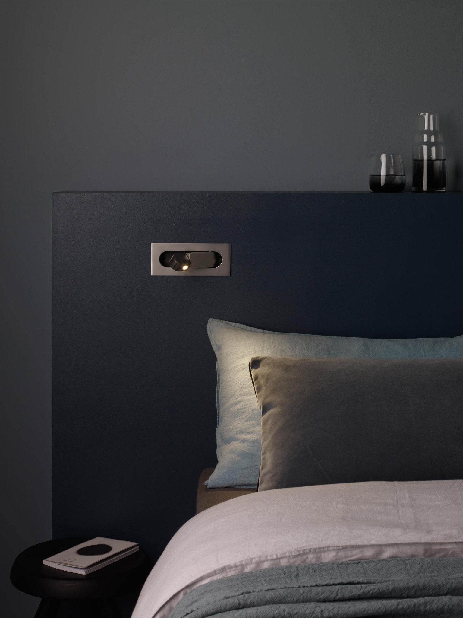 Dark bedroom with a bed and nightstand against a gray wall.