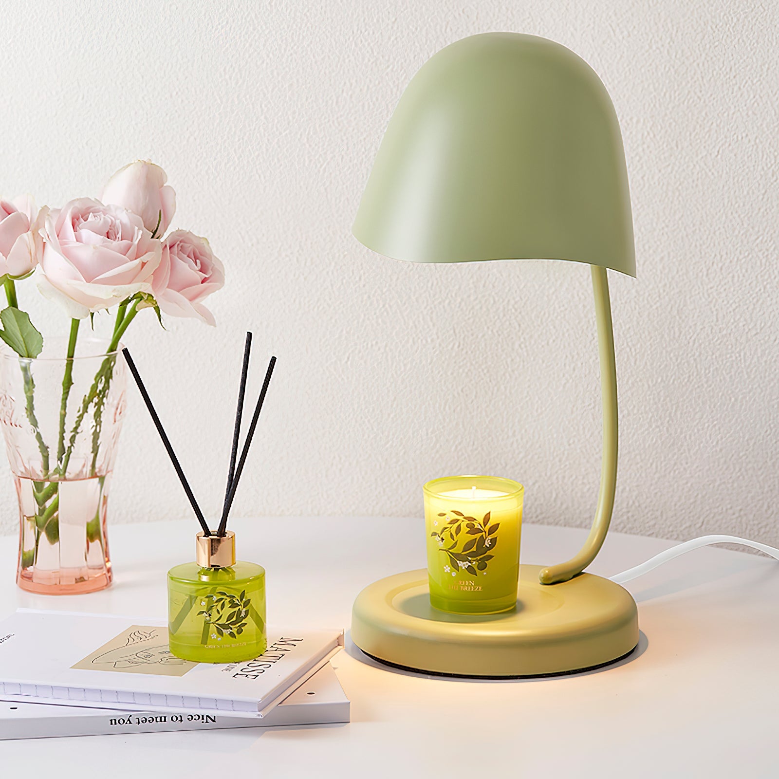 Green table lamp with a candle on a white surface, next to a vase of flowers and a diffuser.