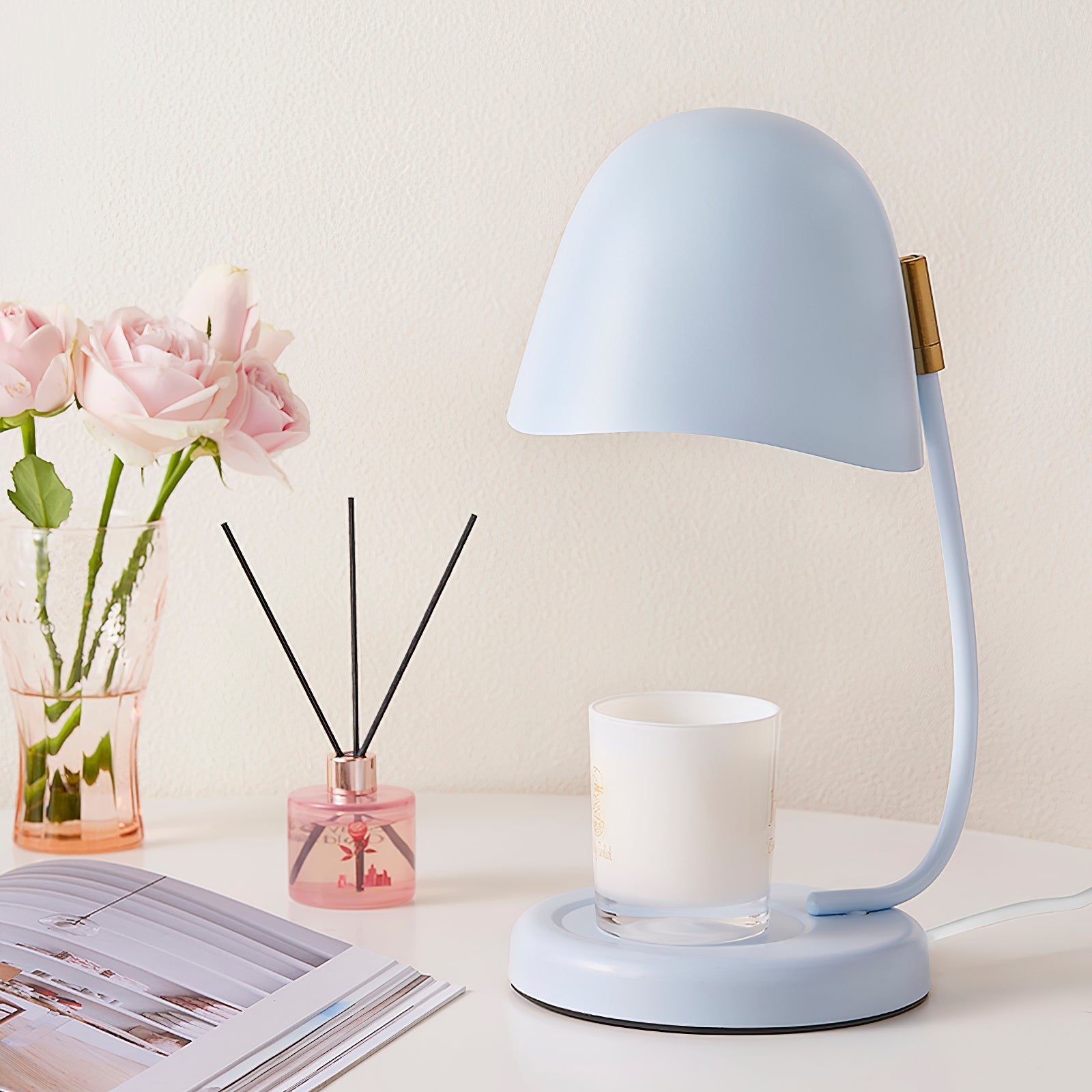 Light blue desk lamp on a table with a magazine, candle, and vase of flowers.